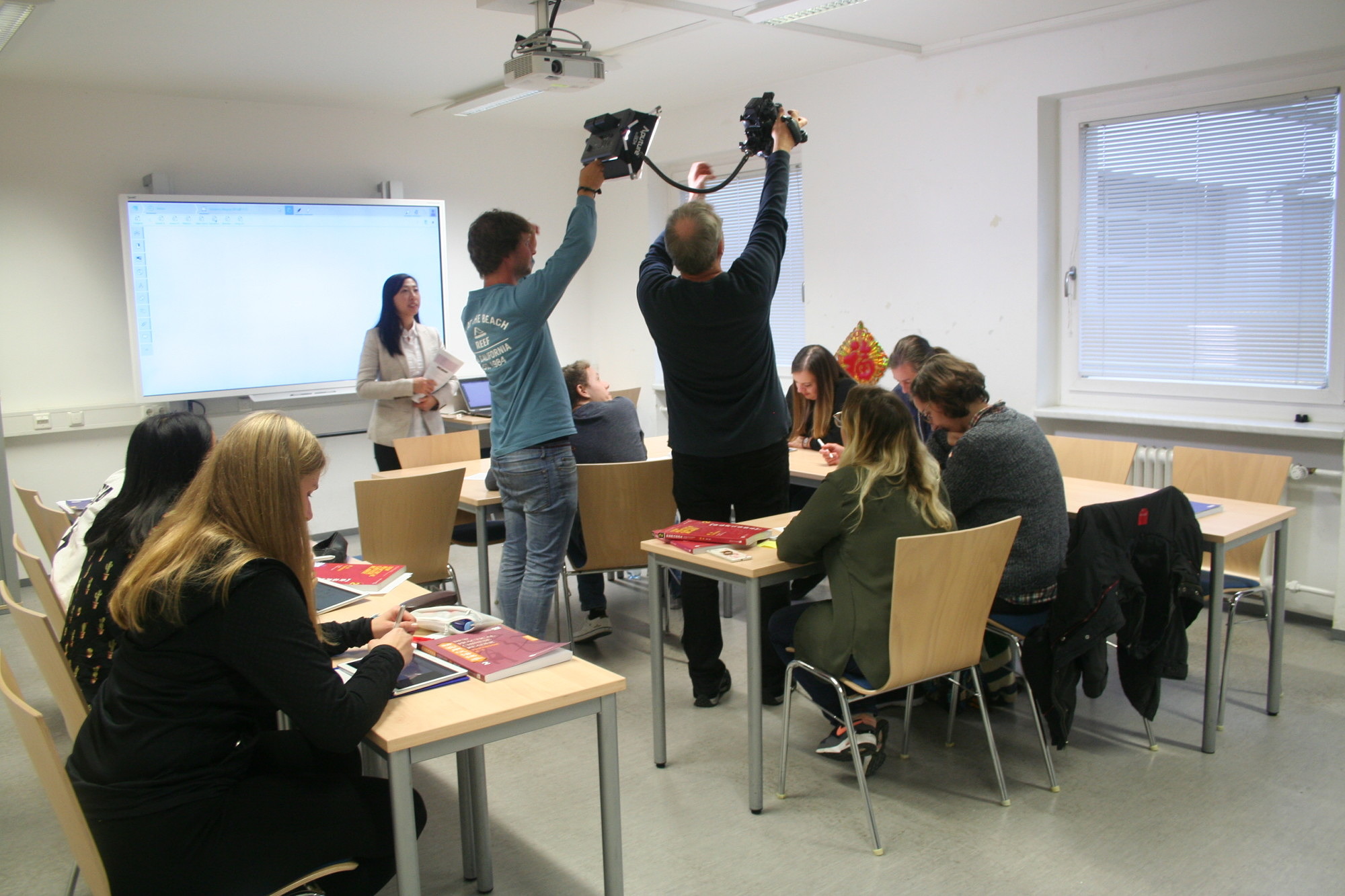 Die Teilnehmer an der Aktion "Filmdreh für das E-learning Projekt" simulieren einen typischen Unterrichtsverlauf mit Tablets, das ganze wird gefilmt. Die Teilnehmer sitzen an Tischen und nutzen Lehrbuch, sowie die Tabletts und schreiben auf diesen u.a. Schriftzeichen.