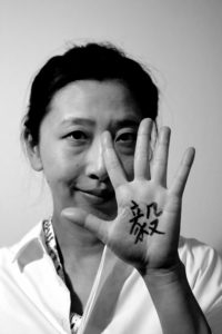 Ein Portraitbild von Frau Cui, welche ihre linke Hand neben ihr Gesicht hält. Auf die Hand wurde das chinesische Schriftzeichen "Yi" geschrieben, welches für "Entschlossenheit" steht.