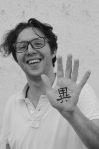 Ein Portraitbild von Herrn Nocchi, welcher seine linke Hand neben sein Gesicht hält. Auf die Hand wurde das chinesische Schriftzeichen "Yi" geschrieben, welches für "Ungewöhnlich" steht.