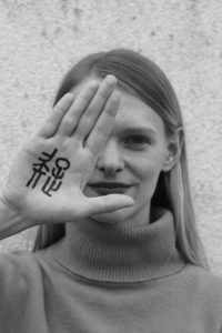 Ein Portraitbild von Frau Schierhorn, welche ihre linke Hand neben ihr Gesicht hält. Auf die Hand wurde das chinesische Schriftzeichen "" geschrieben, welches für "" steht.