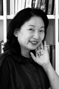 Ein Portraitbild von Frau Dr. Xu-Lackner, welche ihre linke Hand neben ihr Gesicht hält. Auf die Hand wurde das chinesische Schriftzeichen "Heng" geschrieben, welches für "Beständigkeit" steht.