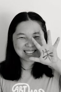 Ein Portraitbild von Frau Zhu, welche ihre linke Hand neben ihr Gesicht hält. Auf die Hand wurde das chinesische Schriftzeichen "Zhu" geschrieben, welches für "Zinoberrot" steht.