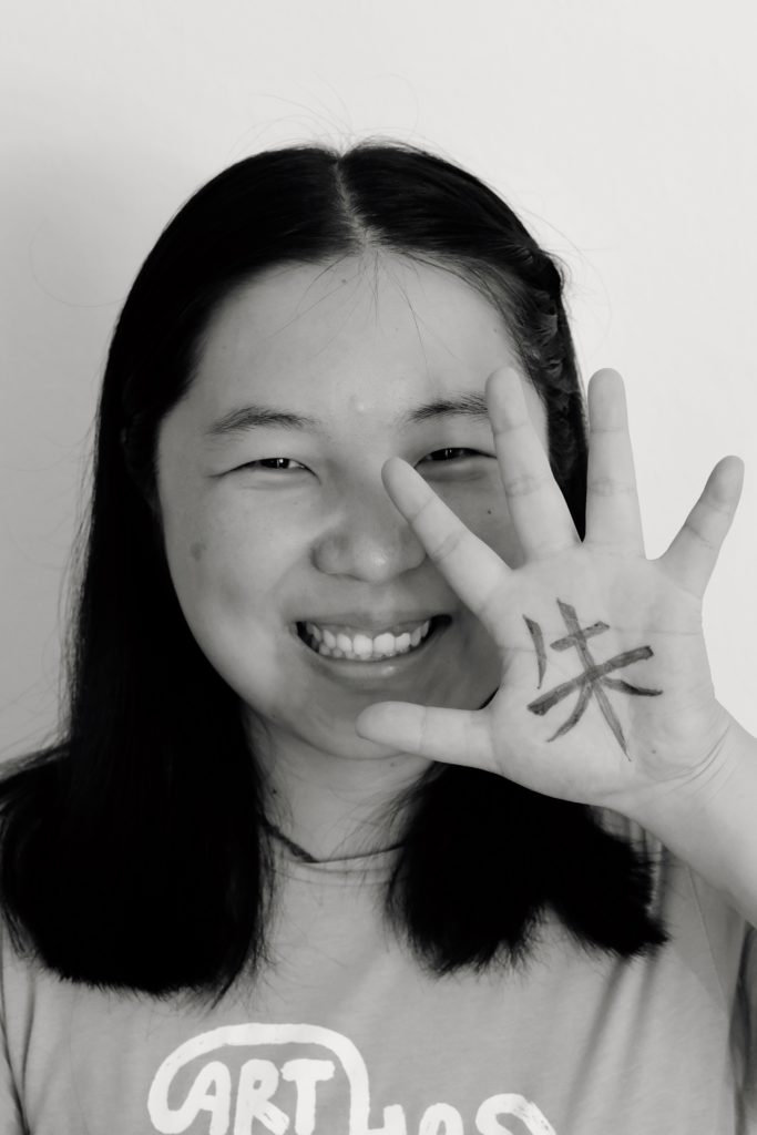 Ein Portraitbild von Frau Zhu, welche ihre linke Hand neben ihr Gesicht hält. Auf die Hand wurde das chinesische Schriftzeichen "Zhu" geschrieben, welches für "Zinnoberrot" steht.