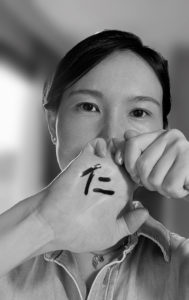Ein Portraitbild von Frau Zhang, welche ihre rechte Hand vor ihr Gesicht hält. Auf die Hand wurde das chinesische Schriftzeichen "Ren" geschrieben, welches für "care and love for the world" steht.